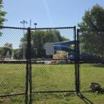 Gate opens to a park playground with a blue slide and yellow supports behind a chain-link fence on a sunny day.