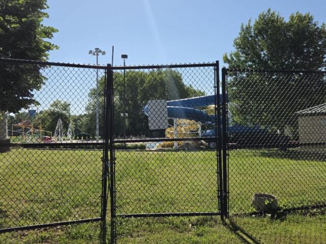 Gate opens to a park playground with a blue slide and yellow supports behind a chain-link fence on a sunny day.