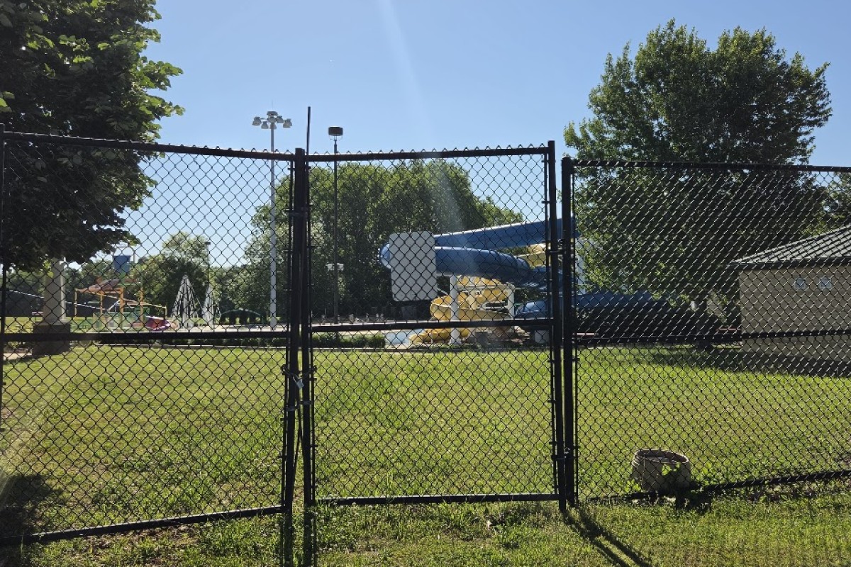 Gate opens to a park playground with a blue slide and yellow supports behind a chain-link fence on a sunny day.