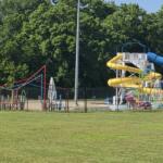 A colorful playground with yellow and blue water slides behind a black fence, set against green trees and a grassy field.