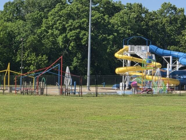 A colorful playground with yellow and blue water slides behind a black fence, set against green trees and a grassy field.