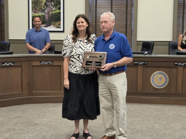 Two adults pose in a government meeting room, holding a wooden award plaque between them as colleagues sit behind at the desk.