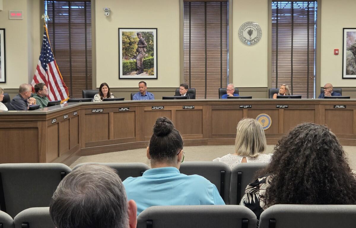 Civic council meeting in a formal chamber with officials seated at a curved wooden dais, American flag nearby and a seal on the wall behind them.
