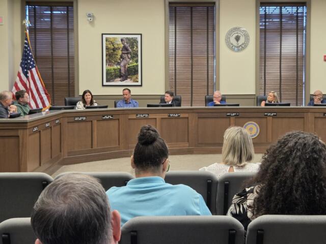 Civic council meeting in a formal chamber with officials seated at a curved wooden dais, American flag nearby and a seal on the wall behind them.