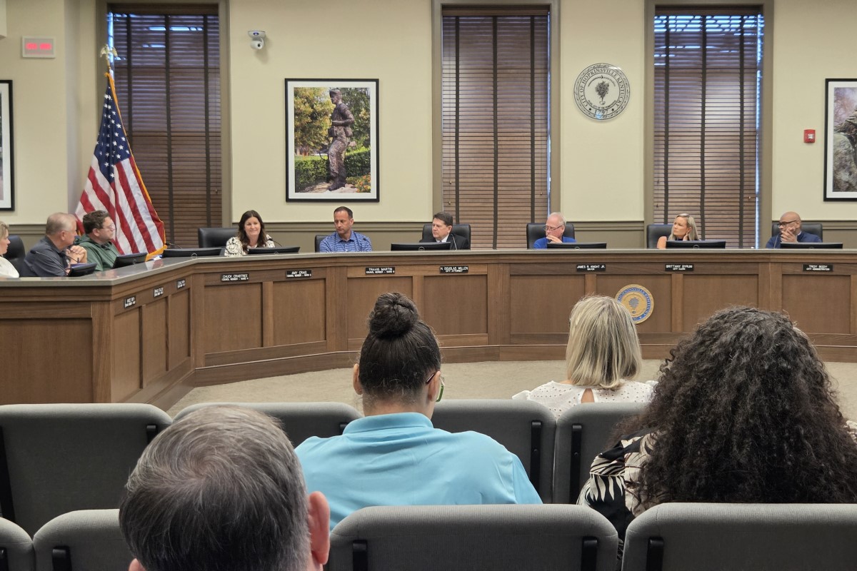 Civic council meeting in a formal chamber with officials seated at a curved wooden dais, American flag nearby and a seal on the wall behind them.