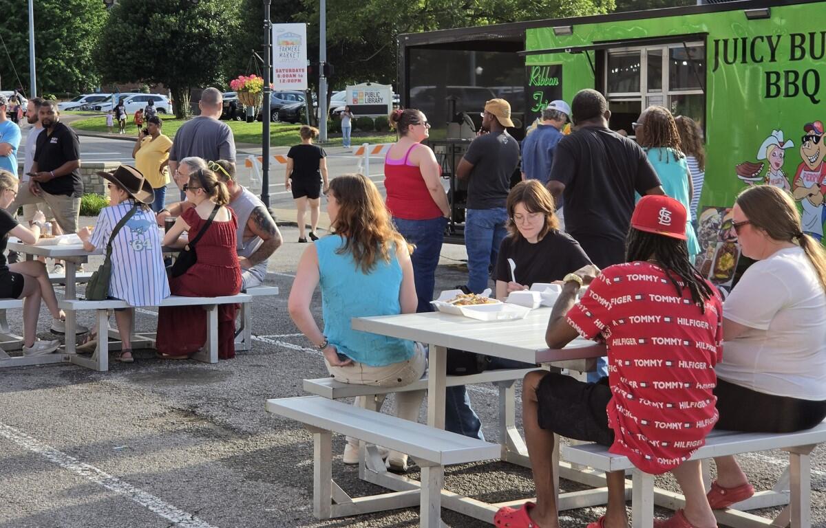 Group of people seated at white picnic tables outside a bright green Juicy BBQ truck, eating and chatting on a sunny day around a community event.