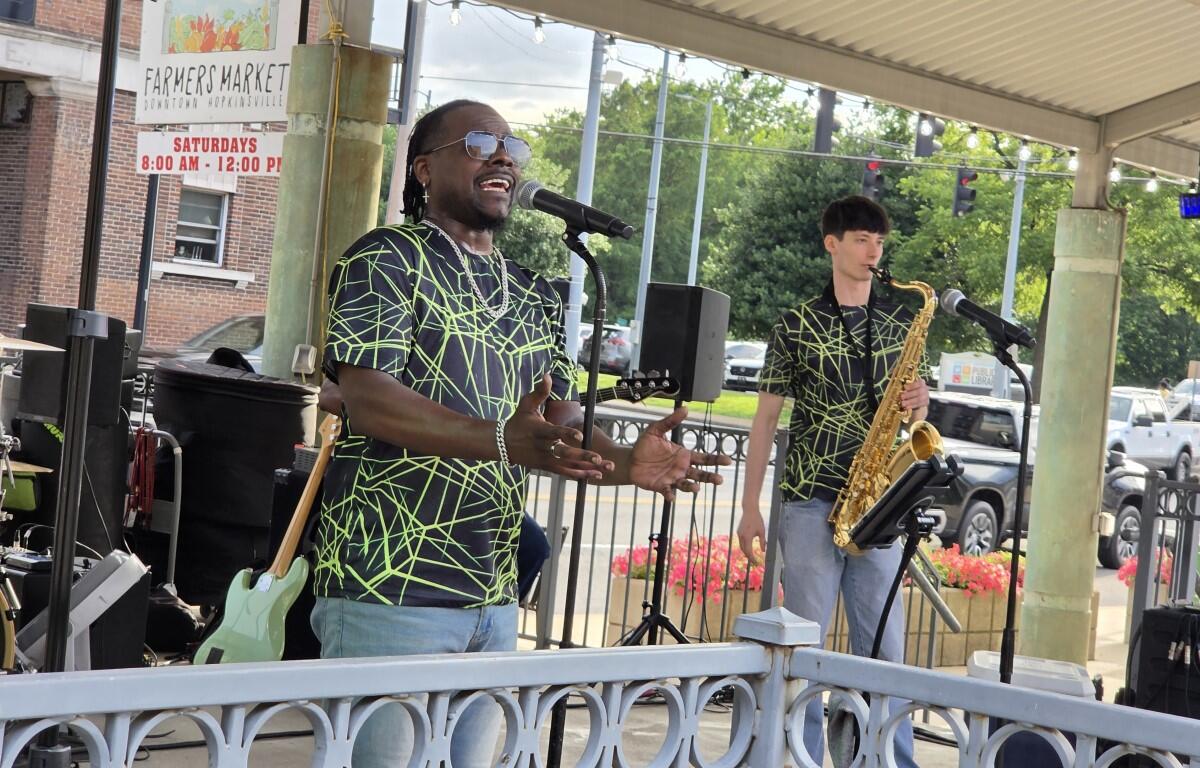 Male singer wearing neon-green web-patterned shirt and sunglasses performs into a microphone on a small outdoor stage; a saxophonist stands nearby with his instrument under a pavilion roof.