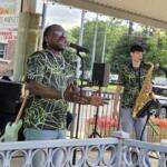 Male singer wearing neon-green web-patterned shirt and sunglasses performs into a microphone on a small outdoor stage; a saxophonist stands nearby with his instrument under a pavilion roof.
