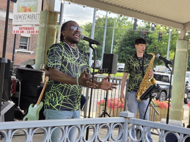 Male singer wearing neon-green web-patterned shirt and sunglasses performs into a microphone on a small outdoor stage; a saxophonist stands nearby with his instrument under a pavilion roof.