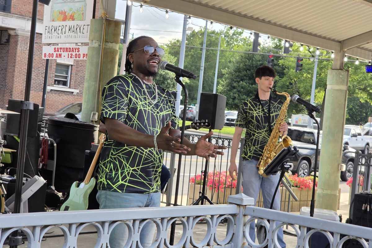 Male singer wearing neon-green web-patterned shirt and sunglasses performs into a microphone on a small outdoor stage; a saxophonist stands nearby with his instrument under a pavilion roof.