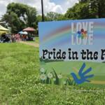Pride in the Park sign with rainbow graphic; park tents and attendees visible in the background while people socialize on the lawn.