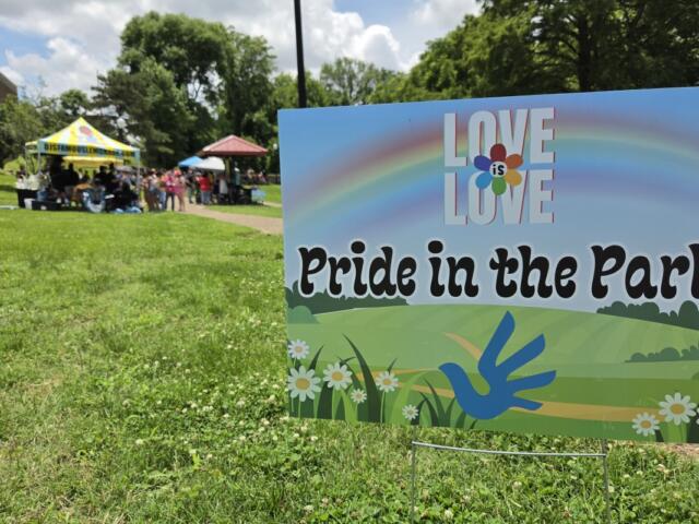 Pride in the Park sign with rainbow graphic; park tents and attendees visible in the background while people socialize on the lawn.