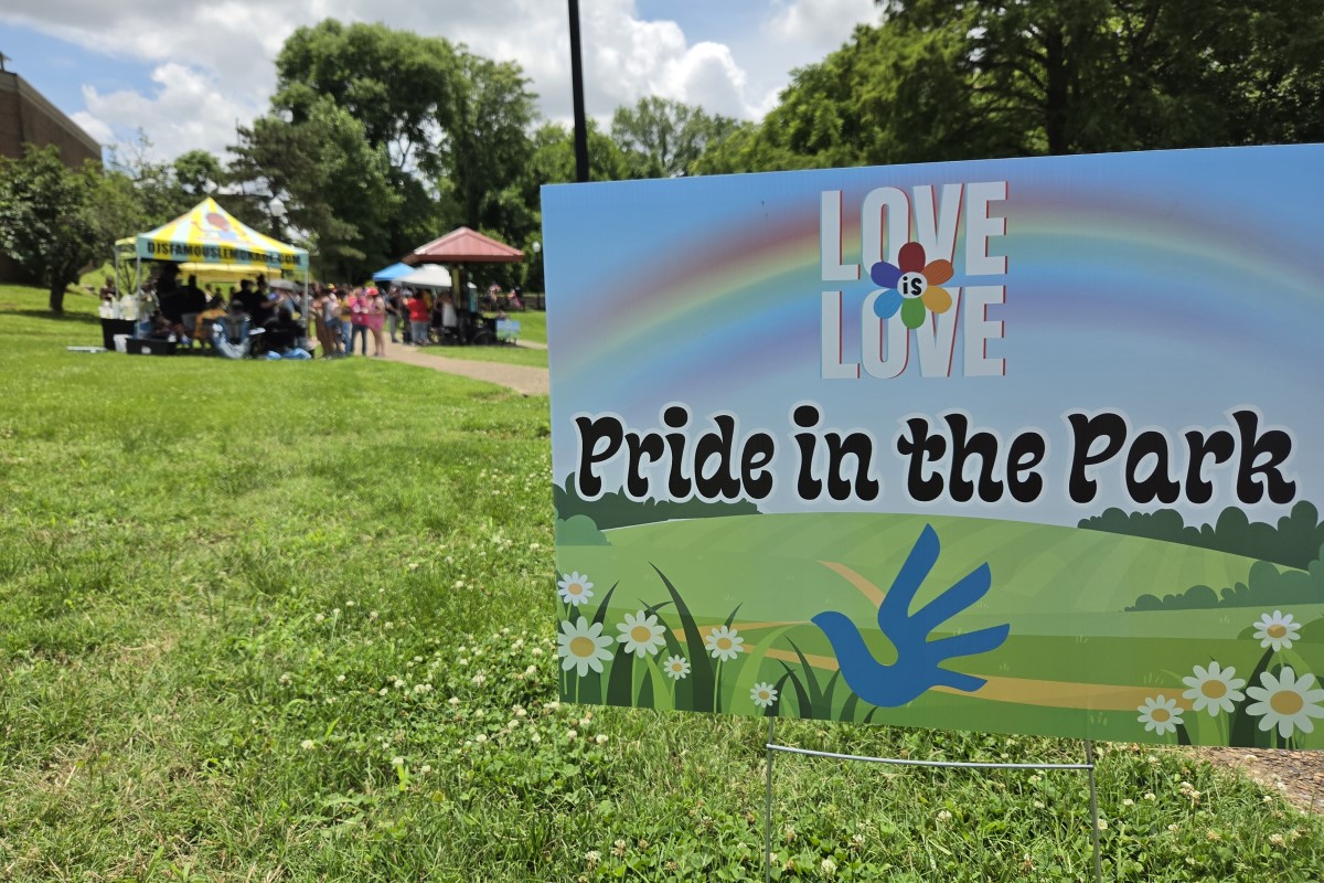 Pride in the Park sign with rainbow graphic; park tents and attendees visible in the background while people socialize on the lawn.