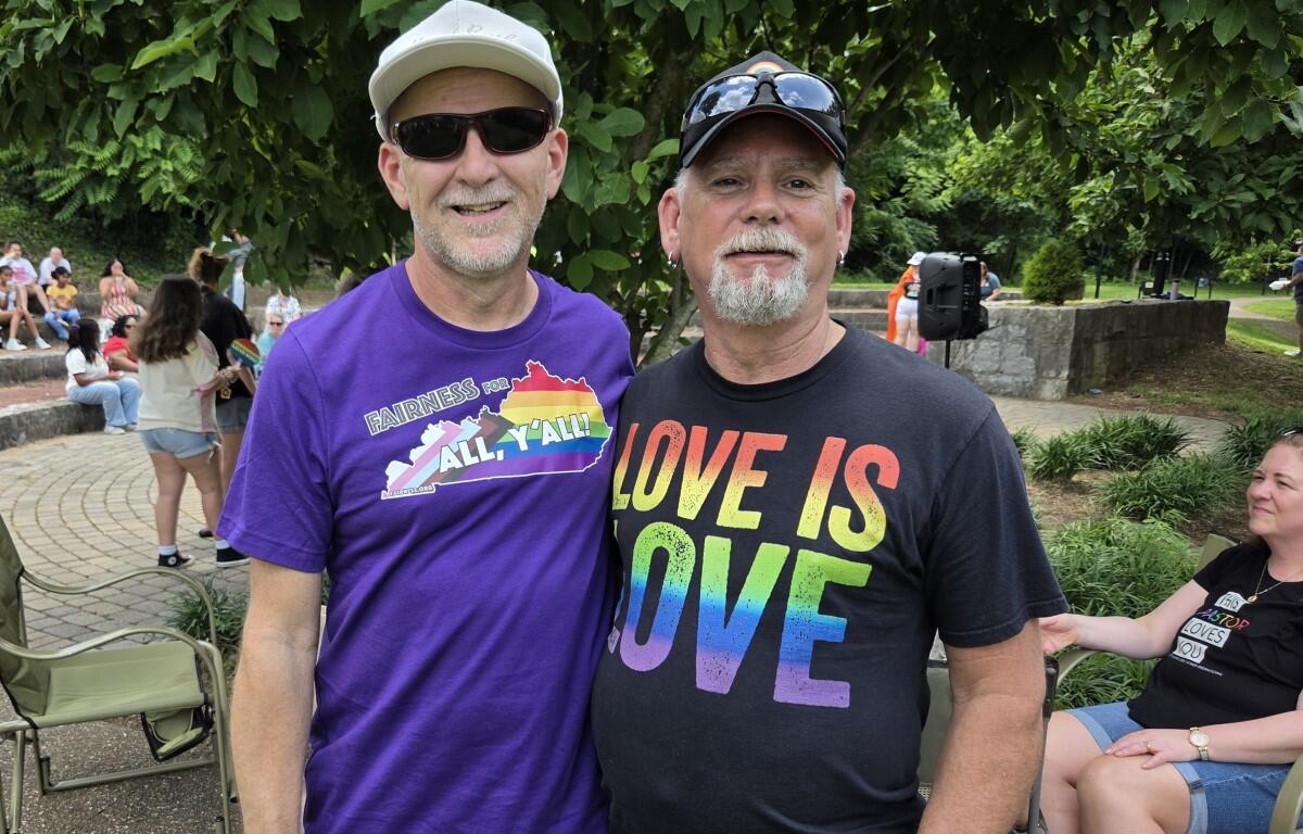 Two men pose together outdoors; one wears a purple 'Fairness for All, Y'all' shirt and white cap, the other a black 'Love is Love' shirt with rainbow colors, smiling.