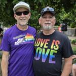 Two men pose together outdoors; one wears a purple 'Fairness for All, Y'all' shirt and white cap, the other a black 'Love is Love' shirt with rainbow colors, smiling.