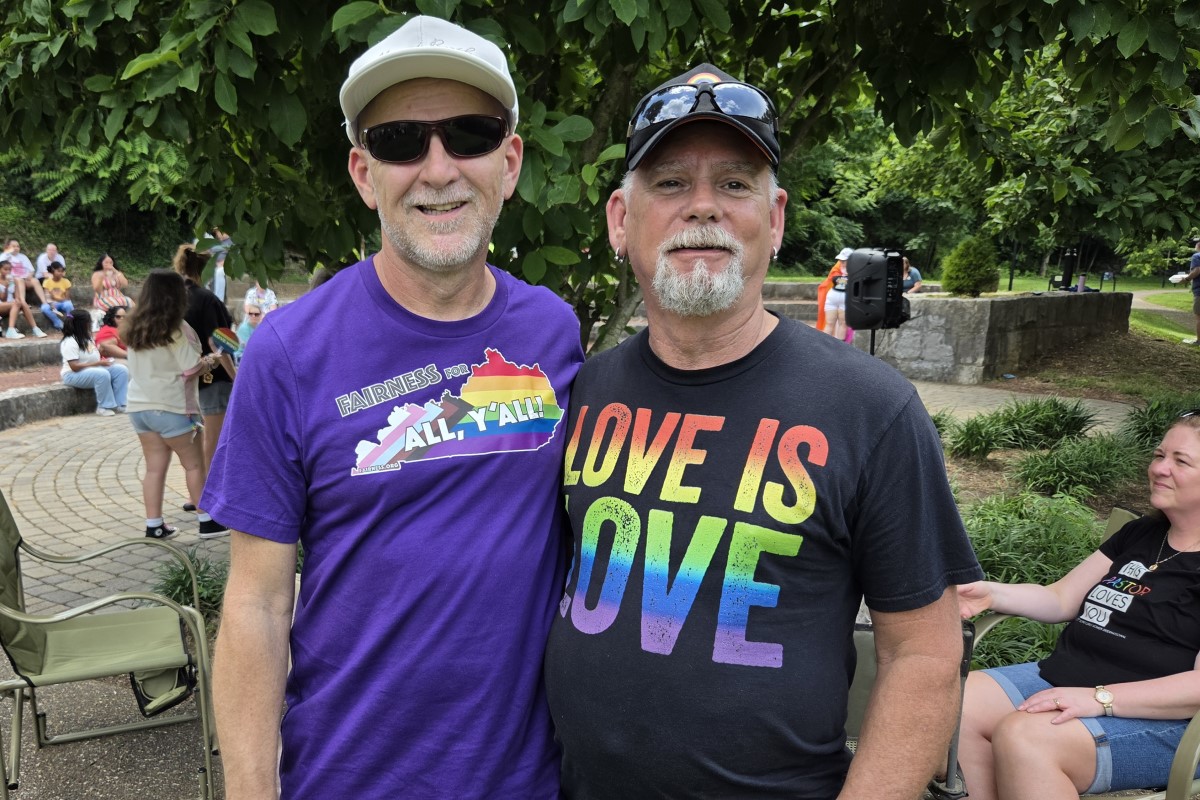 Two men pose together outdoors; one wears a purple 'Fairness for All, Y'all' shirt and white cap, the other a black 'Love is Love' shirt with rainbow colors, smiling.