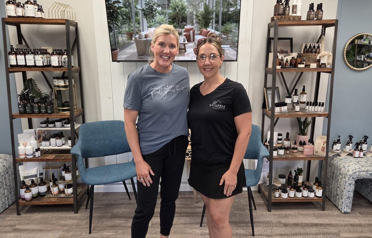 Two women standing in a wellness shop, smiling, with shelves of bottles behind them.