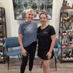 Two women standing in a wellness shop, smiling, with shelves of bottles behind them.