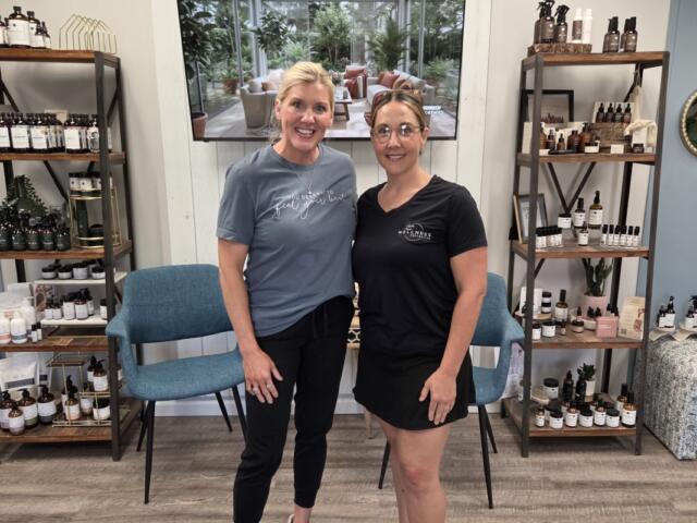 Two women standing in a wellness shop, smiling, with shelves of bottles behind them.
