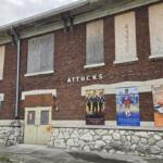Brick building with boarded-up windows and a stone foundation; posters line the lower wall advertising events or shows.