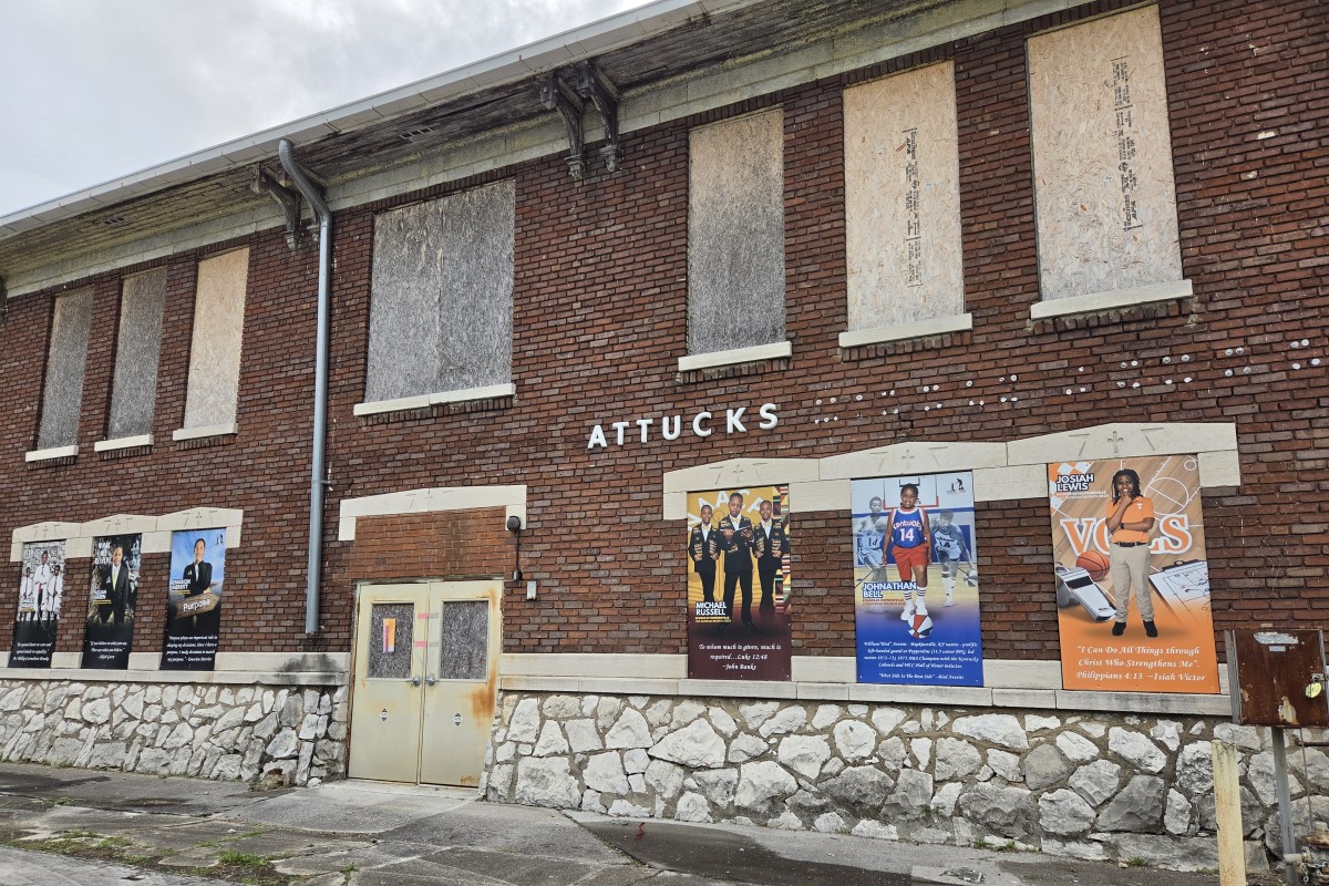 Brick building with boarded-up windows and a stone foundation; posters line the lower wall advertising events or shows.