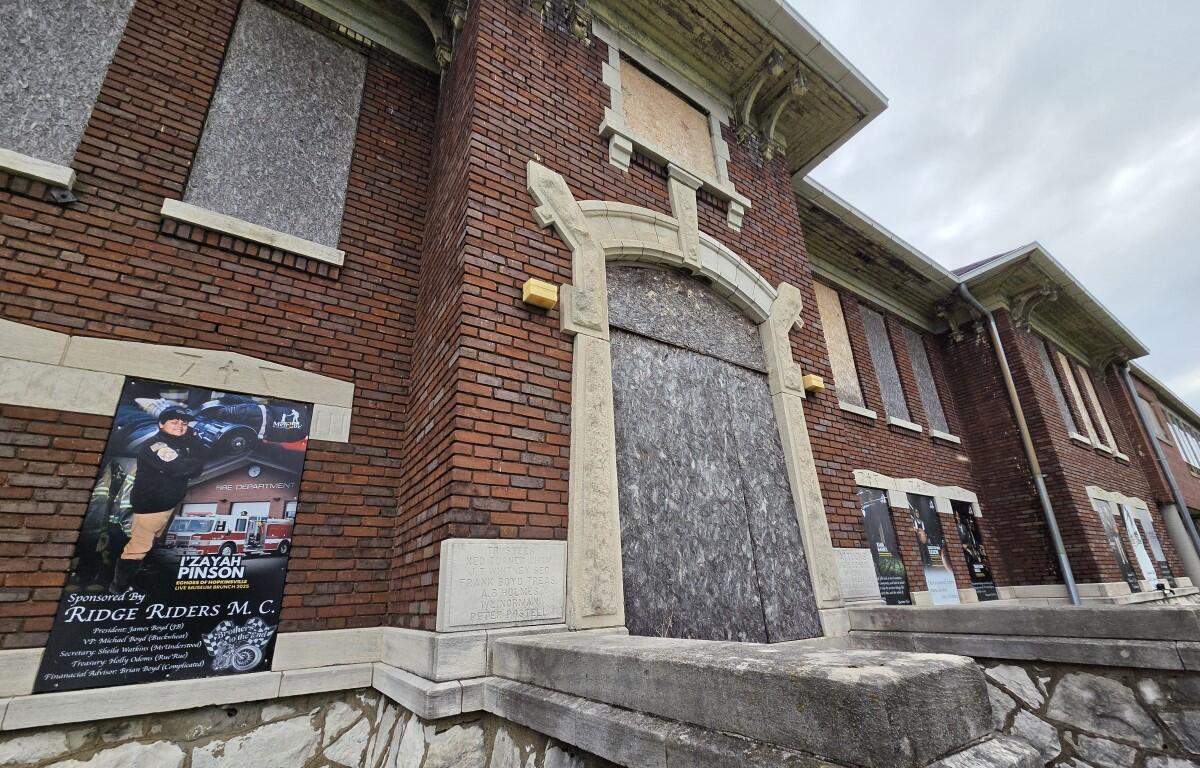 Brick building with a large arched doorway blocked by plywood; multiple posters line the exterior wall to the left and right.