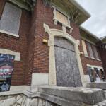 Brick building with a large arched doorway blocked by plywood; multiple posters line the exterior wall to the left and right.