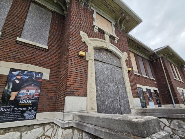 Brick building with a large arched doorway blocked by plywood; multiple posters line the exterior wall to the left and right.
