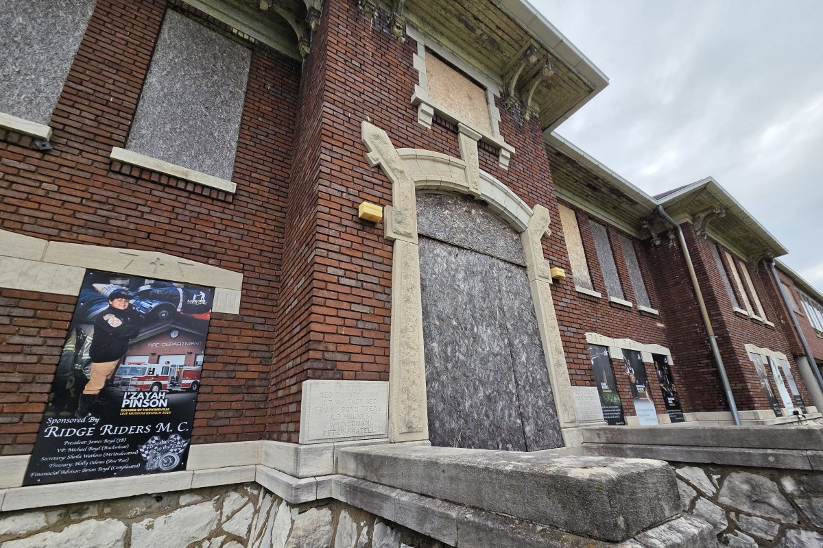 Brick building with a large arched doorway blocked by plywood; multiple posters line the exterior wall to the left and right.