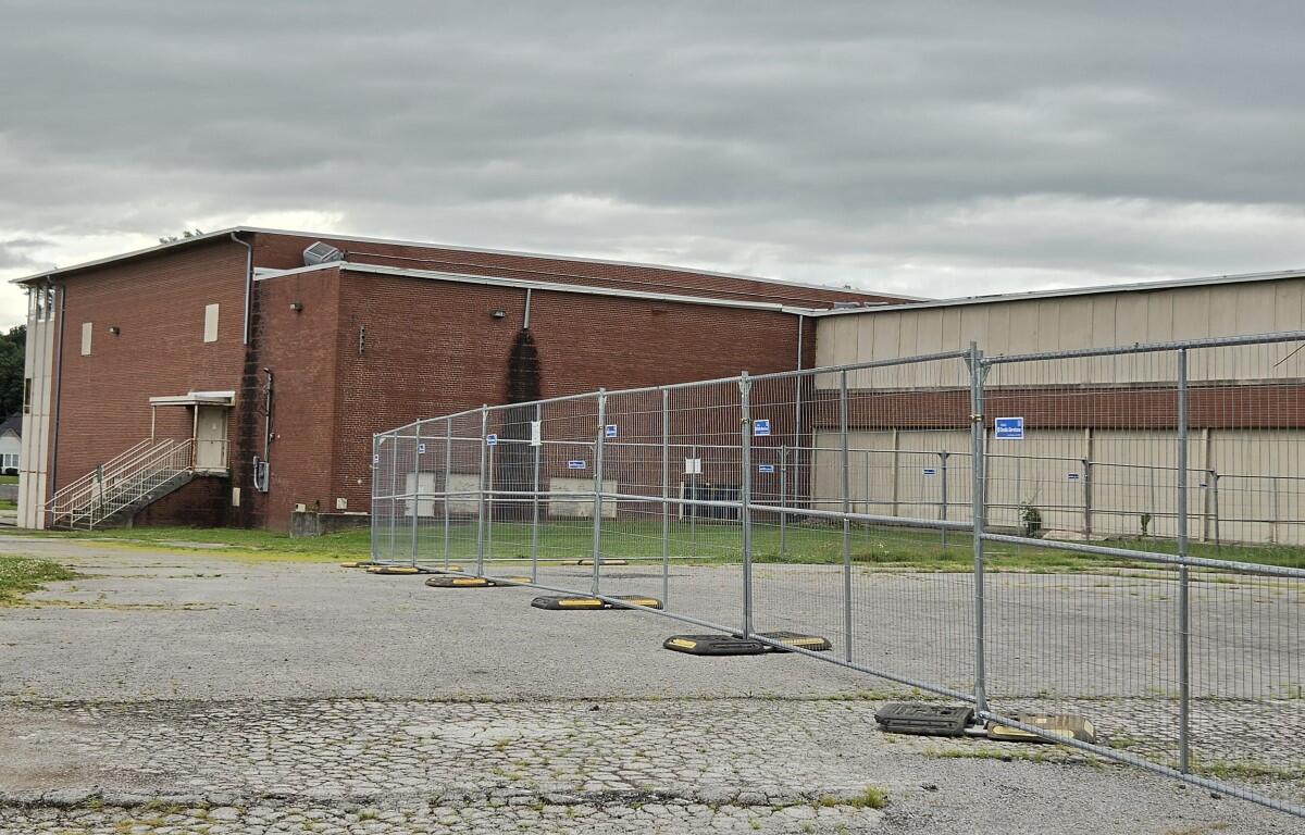 Fenced-off construction area in front of a red brick industrial building under a cloudy sky