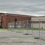Fenced-off construction area in front of a red brick industrial building under a cloudy sky