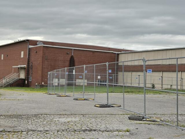 Fenced-off construction area in front of a red brick industrial building under a cloudy sky