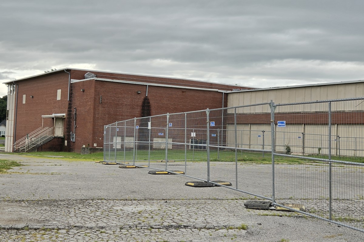 Fenced-off construction area in front of a red brick industrial building under a cloudy sky
