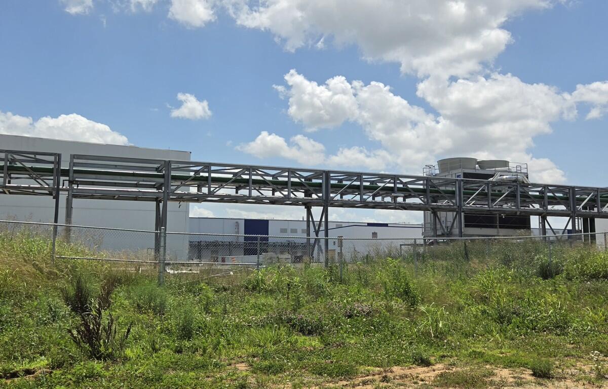 Industrial facility with metal piping elevated on a framework, fenced yard, and overgrown grass under a blue sky with clouds.