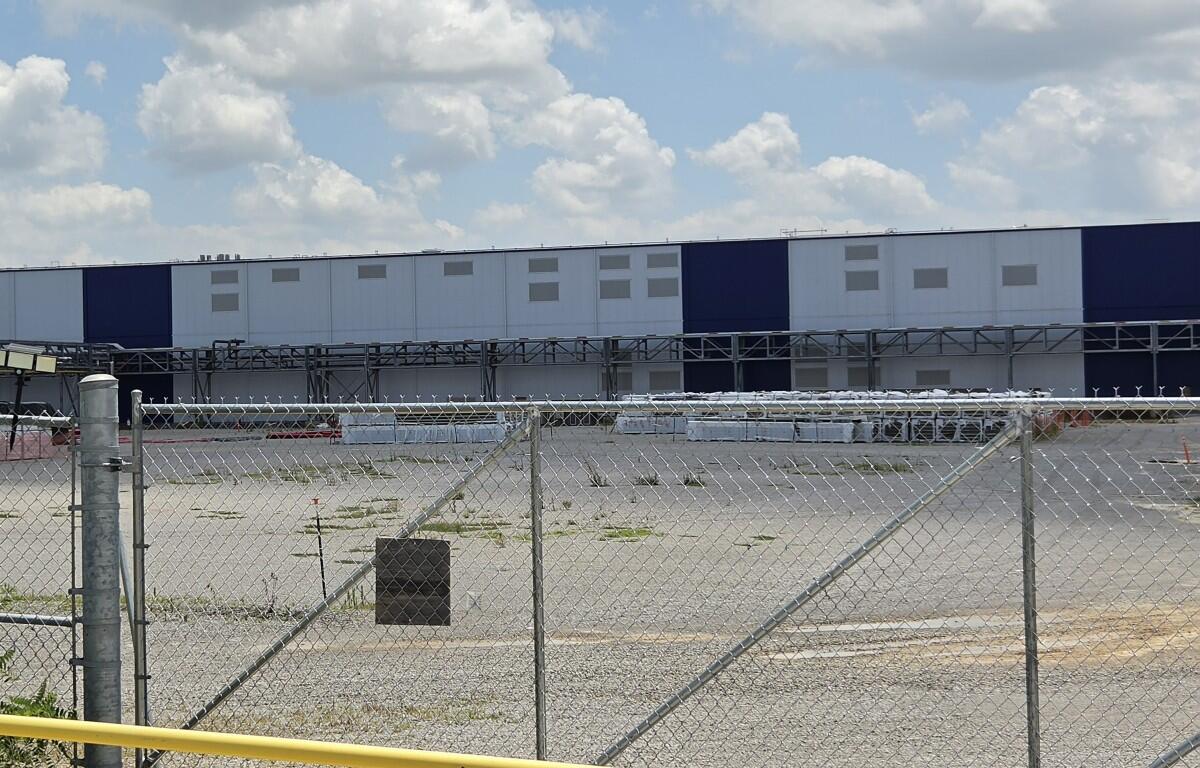 Chain-link fence and gate in foreground with a long blue-and-white industrial warehouse in the background under a cloudy sky.