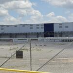 Chain-link fence and gate in foreground with a long blue-and-white industrial warehouse in the background under a cloudy sky.