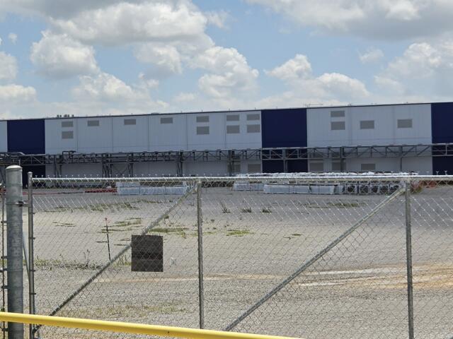 Chain-link fence and gate in foreground with a long blue-and-white industrial warehouse in the background under a cloudy sky.