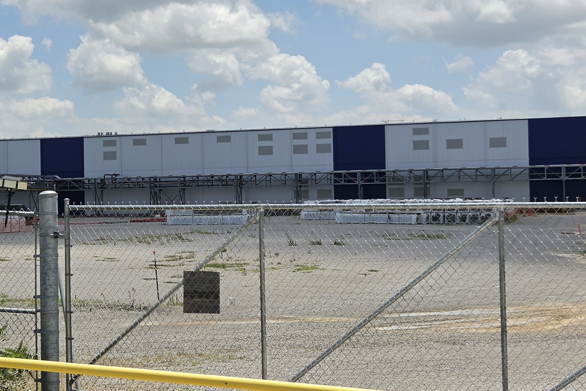Chain-link fence and gate in foreground with a long blue-and-white industrial warehouse in the background under a cloudy sky.