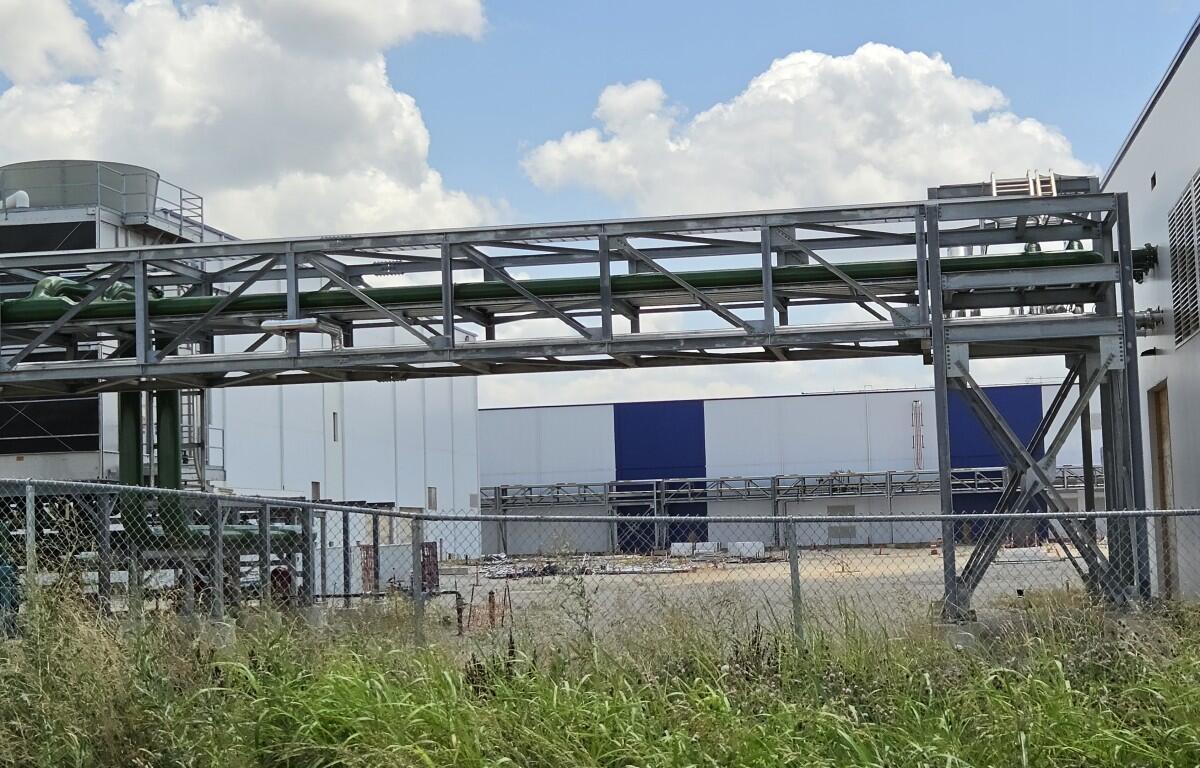 Industrial facility exterior with a metal lattice framework and green pipes spanning above a chain-link fence. Tall grass in the foreground, blue-white warehouse in the background.