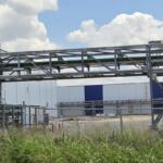 Industrial facility exterior with a metal lattice framework and green pipes spanning above a chain-link fence. Tall grass in the foreground, blue-white warehouse in the background.