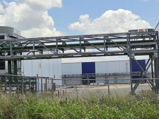 Industrial facility exterior with a metal lattice framework and green pipes spanning above a chain-link fence. Tall grass in the foreground, blue-white warehouse in the background.