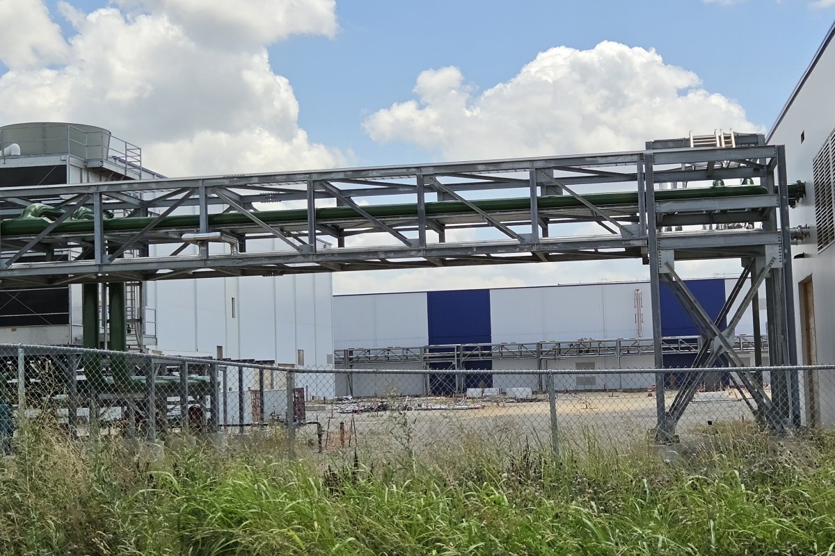 Industrial facility exterior with a metal lattice framework and green pipes spanning above a chain-link fence. Tall grass in the foreground, blue-white warehouse in the background.