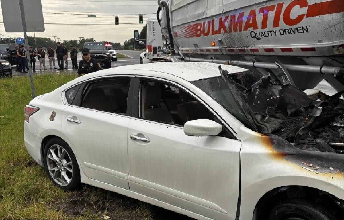 White sedan rear- and side-damaged after colliding with a large truck; front end scorched, shattered windshield, police on scene and bystanders in the background at an intersection.