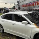 White sedan rear- and side-damaged after colliding with a large truck; front end scorched, shattered windshield, police on scene and bystanders in the background at an intersection.
