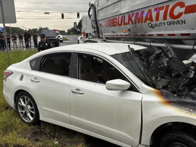 White sedan rear- and side-damaged after colliding with a large truck; front end scorched, shattered windshield, police on scene and bystanders in the background at an intersection.