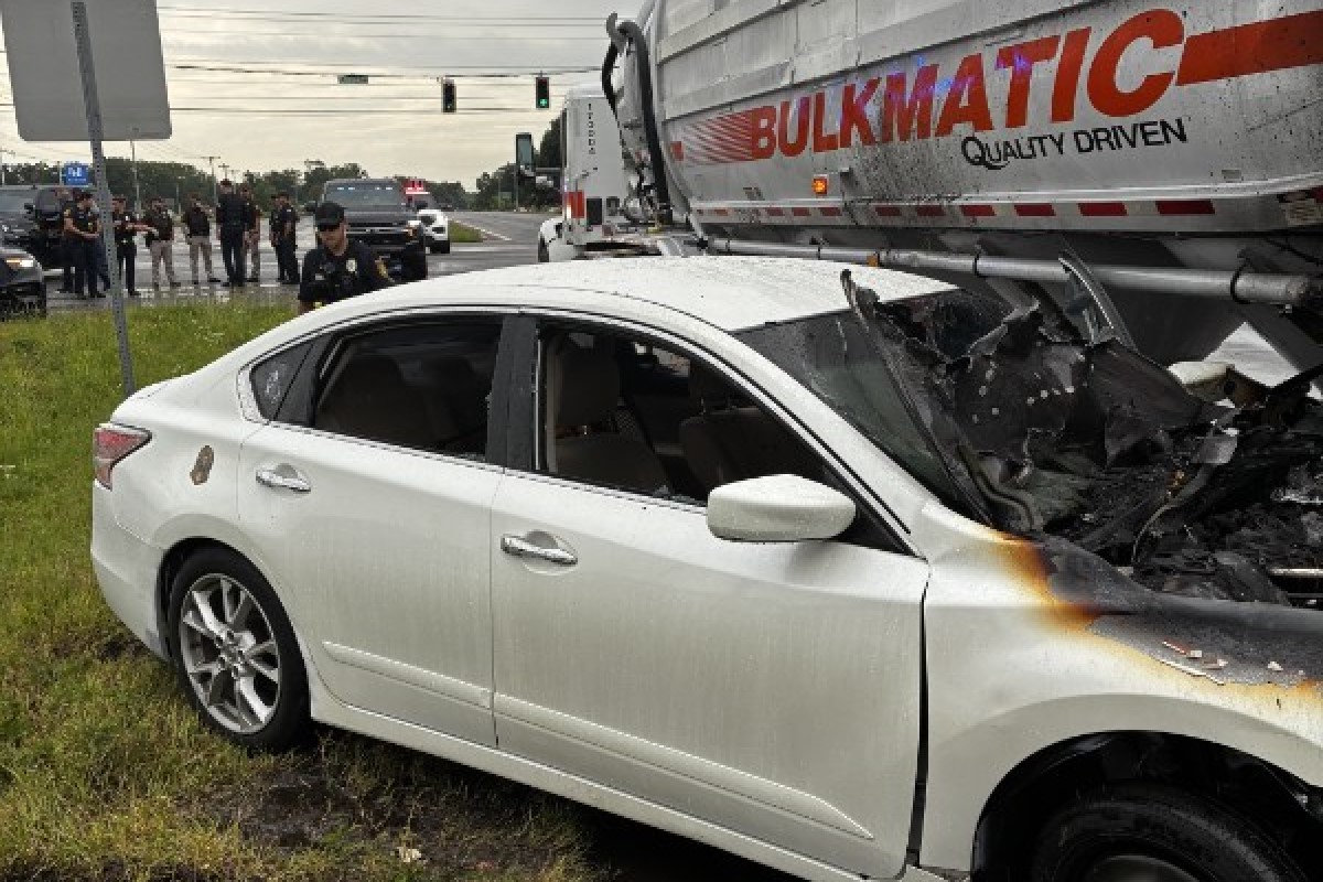 White sedan rear- and side-damaged after colliding with a large truck; front end scorched, shattered windshield, police on scene and bystanders in the background at an intersection.