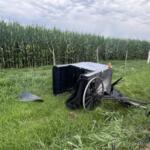Broken horse-drawn wagon with large wheels tipped over beside a grassy roadside near a cornfield fence.