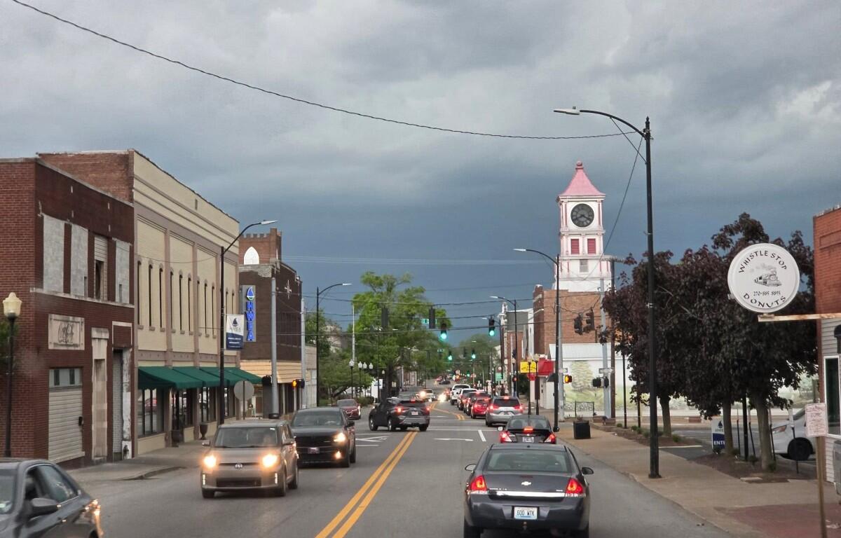 Busy main street with brick storefronts and moving cars, a pink-roofed clock tower visible in the distance under a cloudy sky.