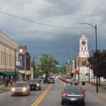 Busy main street with brick storefronts and moving cars, a pink-roofed clock tower visible in the distance under a cloudy sky.