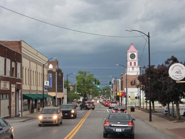 Busy main street with brick storefronts and moving cars, a pink-roofed clock tower visible in the distance under a cloudy sky.